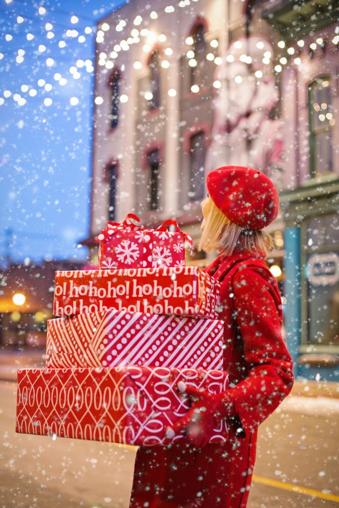 A photo of a person dressed in red walking outside in the snow with a stack of gifts in wrapping paper in their hands. An imaged used to exemplify holiday waste.