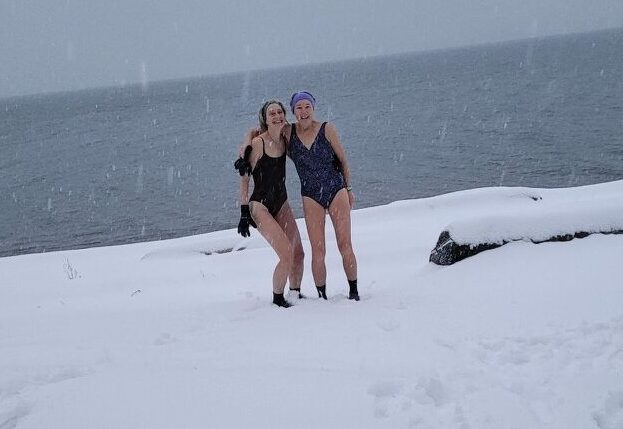 Two smiling people in swimsuits stand on the beach covered in snow. Open Water Swimming is intense!