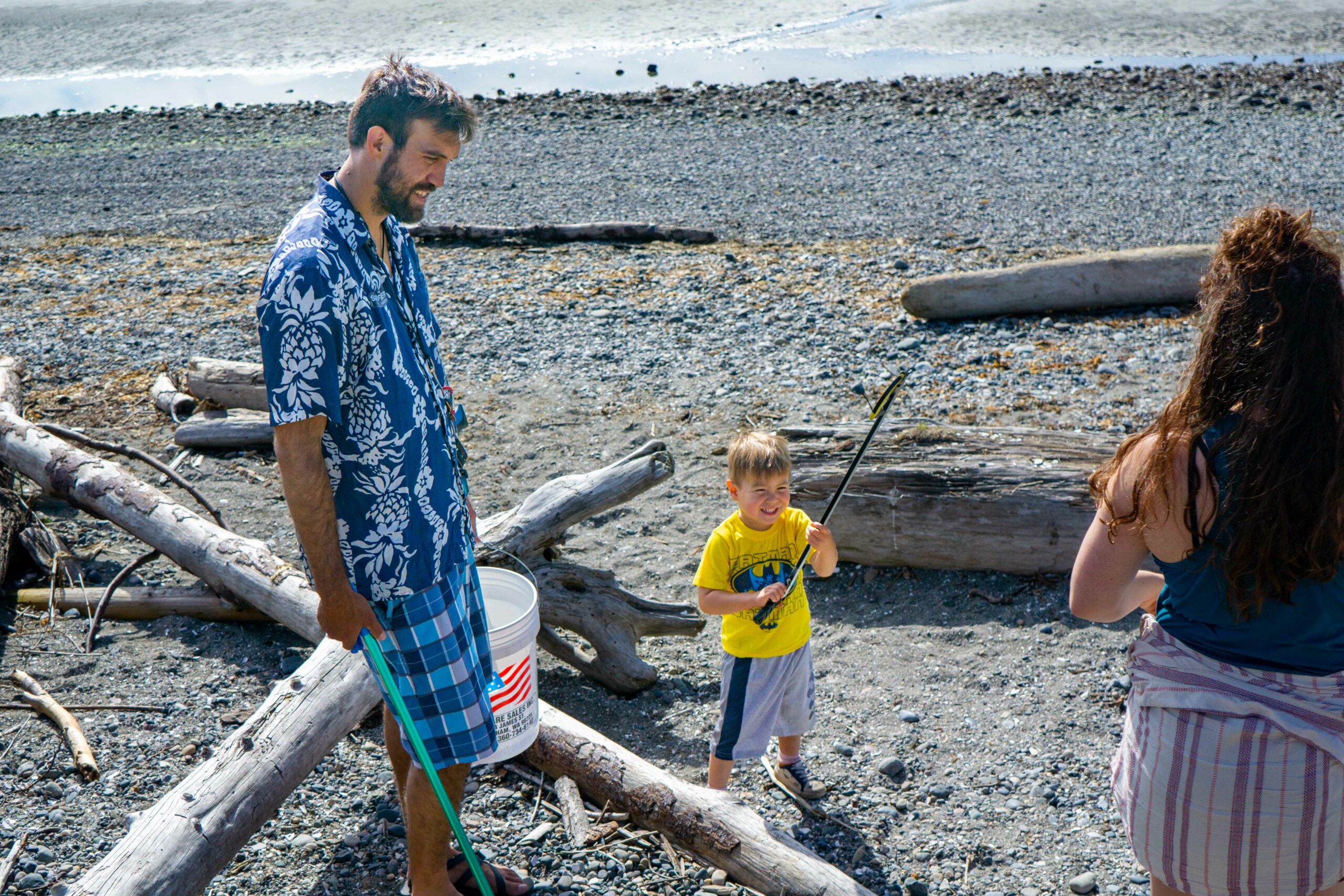 A man and a child holding trash grabbers and buckets on a sandy beach with driftwood